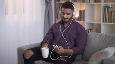 Gadget people. Digital leisure. Modern technology. Relaxed guy listening music in headphones using phone drinking coffee on couch at home interior with free space.