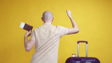 Vacation time. Excited man. Holiday fun. Happy unrecognizable guy with suitcase shaking foreign passport with tickets and dancing on yellow background slow motion.