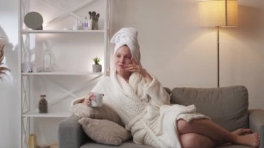 Morning coffee. Weekend relaxation. Sunday home leisure. Happy smiling woman in bathrobe with mug enjoying hot drink daydreaming on couch after bath at light modern interior.