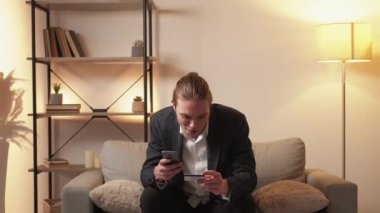 Mobile cashback. Banking app. Successful electronic transaction. Satisfied excited happy business man using credit card phone for virtual money transfer on couch at modern home interior.