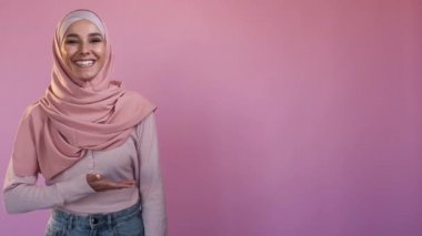 Advertising muslim woman. Modest fashion. Offer recommendation. Happy female in hijab presenting product with hand on pink copy space background.