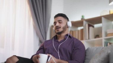 Carefree leisure. Music energy. Home fun. Excited amused man enjoying listening song in headphones singing drinking coffee on couch at light living room interior with free space.