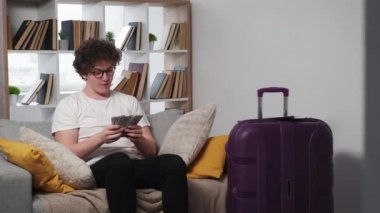 Vacation budget. Excited man. Money calculation. Happy inspired guy sitting sofa with cash dollars packed suitcase ready for travelling light room interior.
