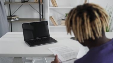 Distance work. Casual man. Online conference. Unrecognizable black guy sitting desk opened laptop blank screen checking documents in light room interior.