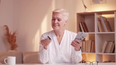 Senior banking. Pension fund. Money transaction. Happy old retired woman choosing dollar banknotes cash credit card payment at home desk.