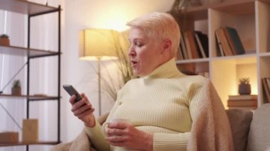 Modern pensioner. Senior mobile leisure. Phone app. Joyful mature woman amazed with smartphone elderly service on couch at cozy home living room.