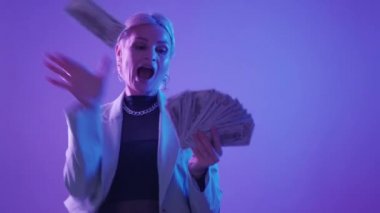 Money rich. Lottery win. Cash millionaire. Happy fancy mature woman throwing dollar banknotes in colorful purple neon light studio.