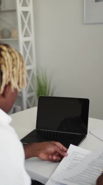 Office work. Concentrated man. Checking documents. Unrecognizable black guy looking at papers and opened laptop blank screen in light room interior vertical.