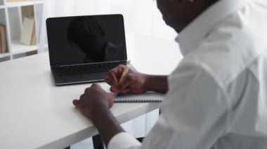 Online education. Studying man. Virtual connection. Elegant black guy sitting work desk with opened laptop dark screen writing in notebook light room interior.