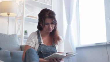 Enjoying reading. Romantic woman. Home relax. Pretty smiling middle-aged lady sitting floor turning pages of book in light room interior.