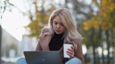 Virtual communication. Happy woman. Distance connection. Pretty smiling lady sitting in autumn park with cup of coffee typing laptop.