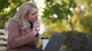 Online meeting. Inspired woman. Virtual connection. Pretty positive lady sitting bench in park talking laptop on coffee break in sunshine day.
