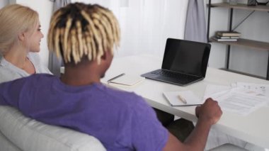 Online education. Casual couple. Video conference. Happy man and woman sitting home desk with copybooks documents looking opened laptop blank screen in light room interior.