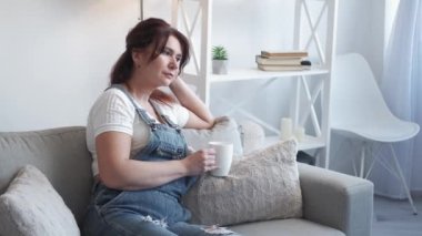 Romantic mood. Dreamful woman. Home relax. Pretty smiling middle-aged lady in casual look sitting sofa with cup coffee in light room interior.