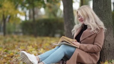 Romantic fall. Happy woman. Enjoying reading. Beautiful smiling lady sitting ground under tree in park with interesting book slow motion.