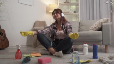 Enjoying music. Meditating woman. Home cleaning. Tranquil adult lady in headphones sitting floor lotus pose surrounding housework staff in light room interior motion view.