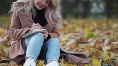 Romantic letter. Inspired woman. Autumn beauty. Pretty smiling lady sitting on leaves ground making notes in notebook.