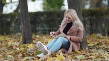 Enjoying reading. Inspired woman. Beautiful autumn. Pretty smiling lady sitting on ground leaning tree in park with interesting book slow motion.