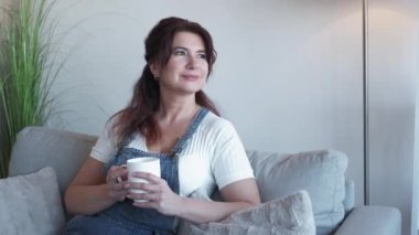 Pleasant day. Pretty aged woman. Coffee break. Dreamful smiling lady in casual look sitting sofa with cup of hot beverage light room interior.