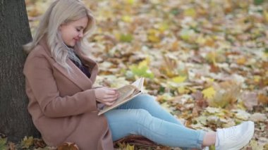 Enjoying reading. Smiling woman. Romantic fall. Pretty happy lady sitting on leaves ground leaning tree with interesting book slow motion.