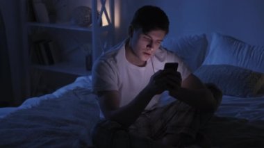 Late mobile chat. Internet fatigue. Sleep disorder. Exhausted tired yawning guy nodding using phone texting message on bed in dark night bedroom.