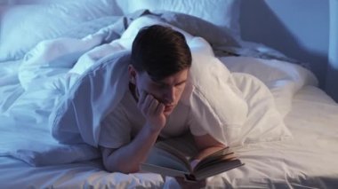 Reading in bed. Night literature. Bedtime leisure habit. Tired curios sleepy man nodding with open book under blanket late in dark bedroom.