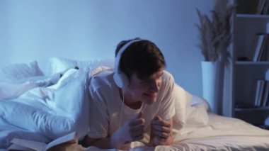 Music in bed. Carefree home evening. Positive playlist. Happy inspired relaxed guy dancing enjoying rythm in headphones in dark bedroom.
