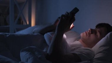 Night chat. Internet fatigue. Social media addiction. Tired exhausted bored man falling asleep using phone in bed late in dark bedroom.