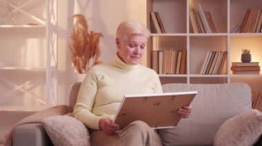 Nostalgia memories. Remembering past. Family missing. Dreamy senior woman pensioner holding looking at photo on couch at home living room.