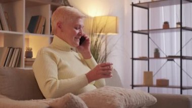 Mature woman call. Phone talk. Grandparent communication. Cheerful grandmother with coffee enjoying smartphone conversation relaxing on couch at home living room.