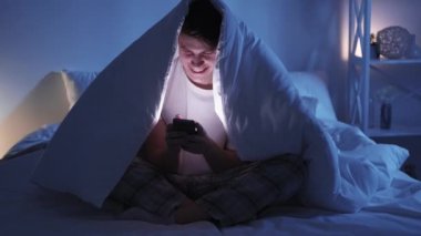 Bed chat. Night online. Late digital leisure. Curious smiling guy using smartphone enjoying fun virtual conversation under blanket in dark bedroom.