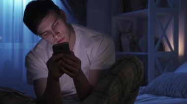 Mobile fatigue. Internet insomnia. Social media addiction. Exhausted bored man falling asleep using phone late on bed in dark night bedroom.