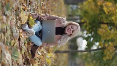 Virtual conference. Happy woman. Romantic autumn. Pretty smiling lady sitting on leaves ground talking on laptop in park vertical.