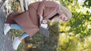 Virtual connection. Positive woman. Digital life. Beautiful happy lady sitting bench in park talking laptop holding cup of coffee vertical.