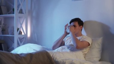 Bedtime tea. Cozy home evening. Weekend relaxation. Inspired happy smiling man enjoying hot herbal drink in bed in dark modern bedroom with free space.