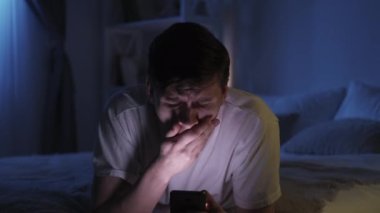 Online insomnia. Gadget night. Mobile overuse. Tired bored sleepy yawning guy using phone surfing Internet chatting late on bed in dark bedroom.