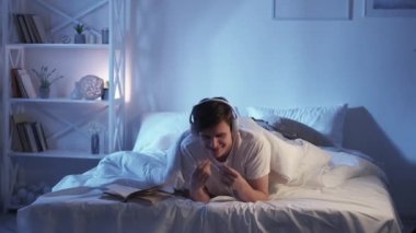 Home music leisure. Night inspiration. Positive vibe. Happy amused relaxed guy enjoying listening to playlist in headphones alone in bed in modern bedroom with free space.