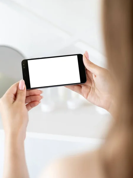 Online connection. Mobile mockup. Digital life. Unrecognizable woman holding smartphone with blank screen in light bathroom interior blur.