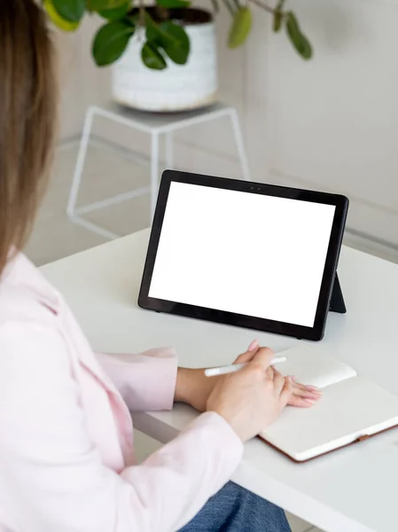 Online conference. Studying woman. Computer mockup. Unrecognizable lady looking tablet computer blank screen sitting desk with copybook in light room interior.