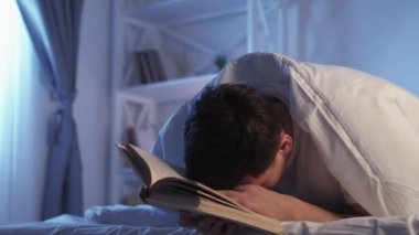 Night reading. Learning fatigue. Late study. Tired exhausted student guy nodding falling asleep with open book under blanket in bed.