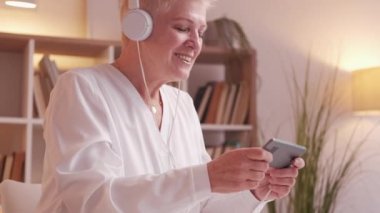 Modern senior. Gadget leisure. Retirement fun. Happy carefree mature woman pensioner playing mobile phone game in headphones at home.
