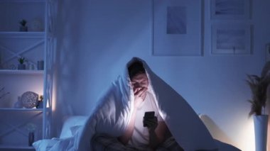 Mobile fatigue. Gadget insomnia. Social media addiction. Exhausted bored sleepy yawning man using phone chatting late under blanket on bed in dark night bedroom.