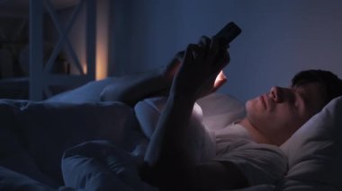 Night chat. Internet fatigue. Social media addiction. Tired exhausted bored man falling asleep using phone in bed late in dark bedroom.