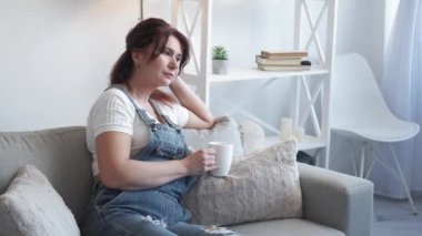 Romantic mood. Dreamful woman. Home relax. Pretty smiling middle-aged lady in casual look sitting sofa with cup coffee in light room interior.