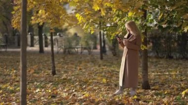 Romantic mood. Pretty woman. Mobile connection. Beautiful smiling lady staying in autumn park with cup of coffee typing smartphone.