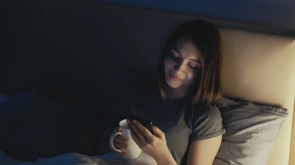 Vertical video. Online connection. Sleepless woman. Pretty lady in bed scrolling social media in smartphone drinking tea in light shadow room interior.