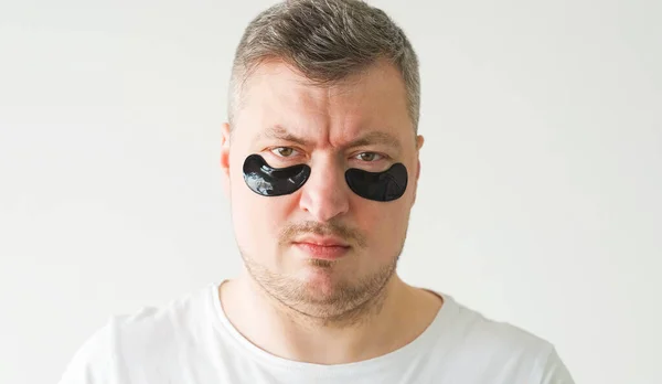 Vertical video. Confused man. Face treatment. Anti-aging procedure. Dissatisfied gloomy guy with hydrogel black under eye patches on light background.