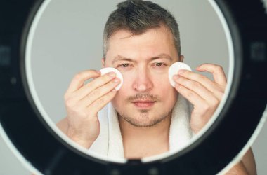 Dikey video. Erkek cilt bakımı. Temizleme prosedürü. Yüz bakımı. Kendine güveni tam yakışıklı bir adam. Gri arka plan lambası üzerine pamuk pedlerle tonik sürüyor..