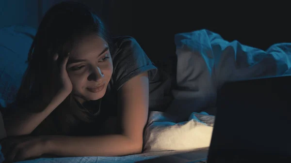 垂直ビデオ オンライン映画 眠れない女だ 夜のレジャー 可愛いです笑顔の女性寝そべってベッド見てオープンラップトップで暗い部屋のインテリア — ストック写真