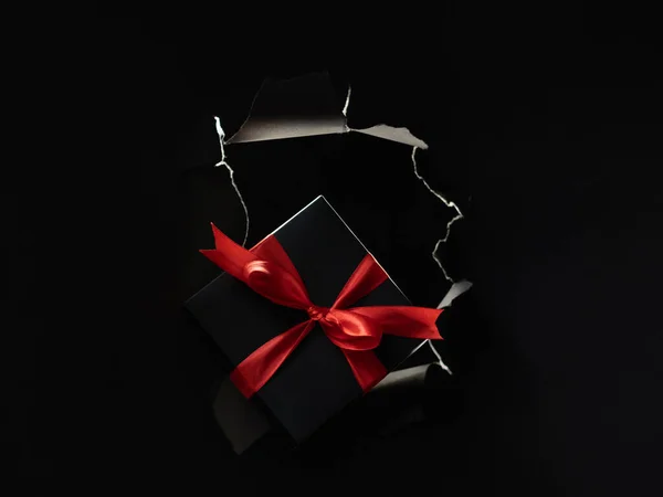 Black Friday sale. Holiday present. Night shopping. Gift box with red ribbon in breakthrough paper hole ripped wall isolated on dark copy space advertising background.